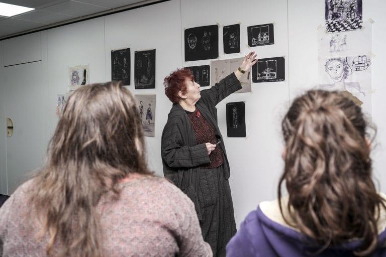 Pamela Howard points to some pictures on a wall in the middle of a lecture. Two students are viewed from behind watching the lecture.
