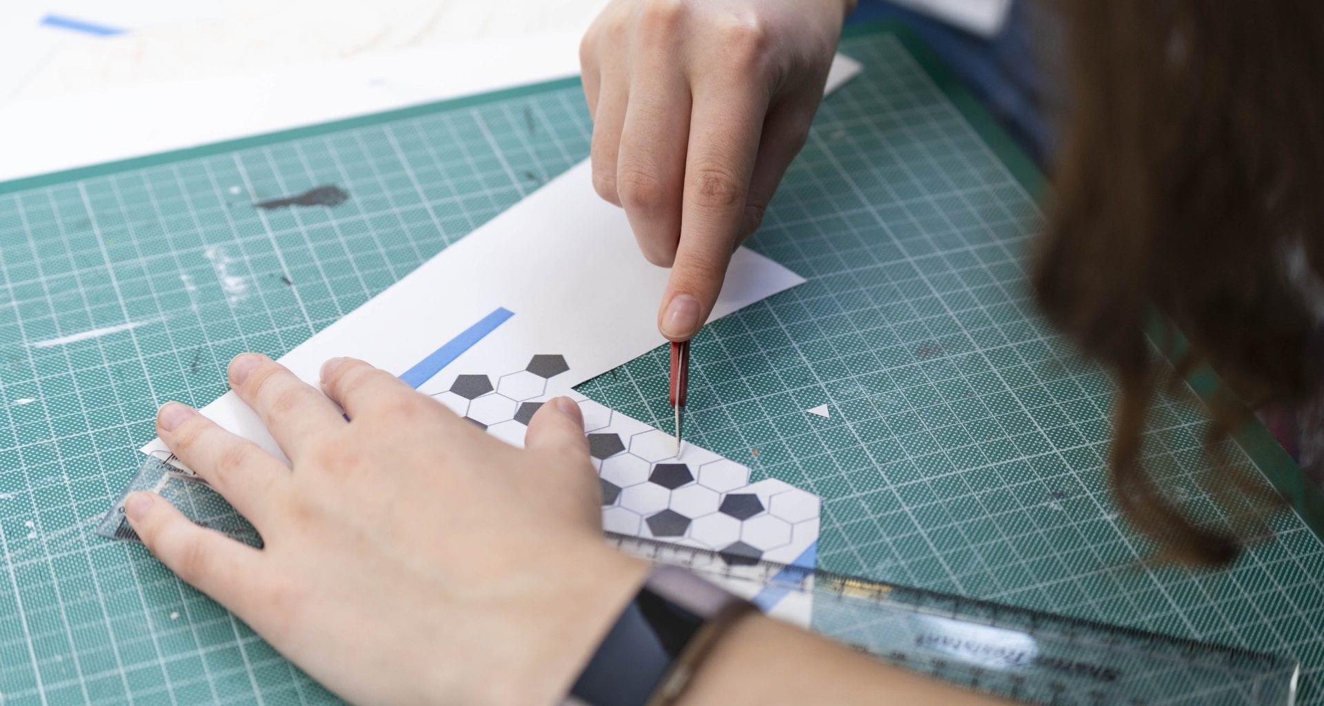 A person uses a cutting tool on a piece of card with hexagon shapes on it, leaning on a cutting board.