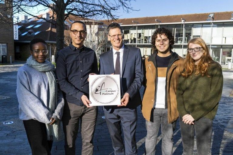 Five people standing in a courtyard posing for a photo with an award. The award says "Eco Campus Platinum".