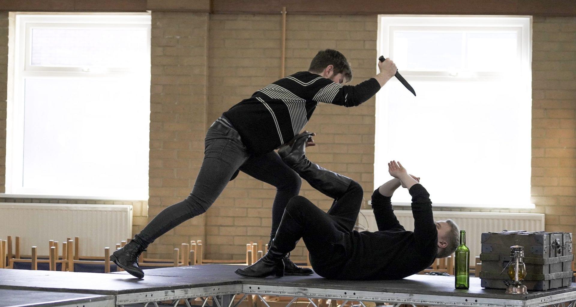 Two actors on a stage, one standing and leaning over holding a knife, the other on their back with a leg in the air and their arms in front of their face.