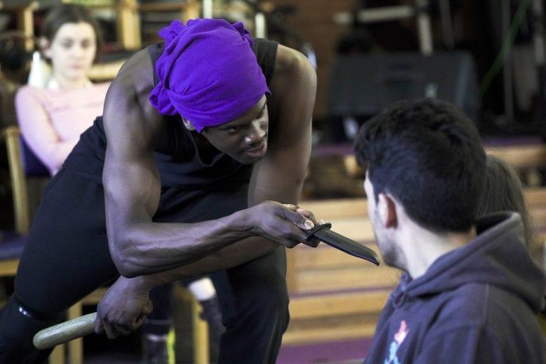 A person wearing a bandana leans over and points a knife at another person sitting in front of them.