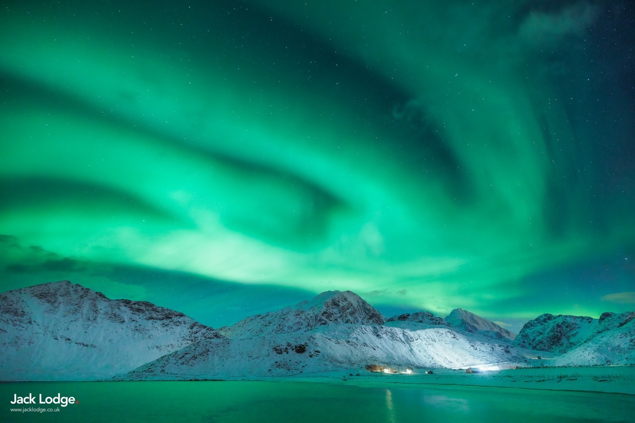 A large aurora sweeping the sky above snowy mountains.