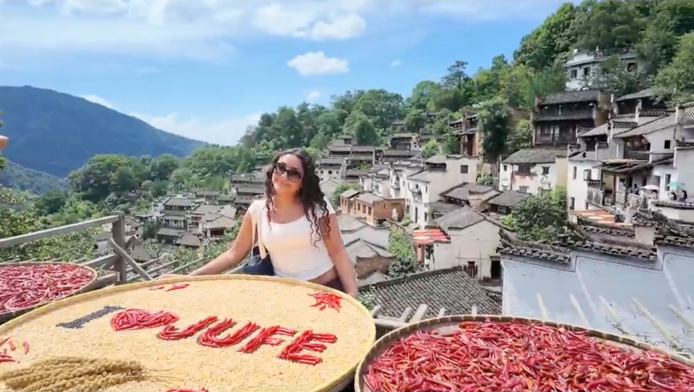 A woman in sunglasses sits beside large trays of chilli peppers and a woven display spelling 'MUFU', with a hillside village and green mountains in the background.