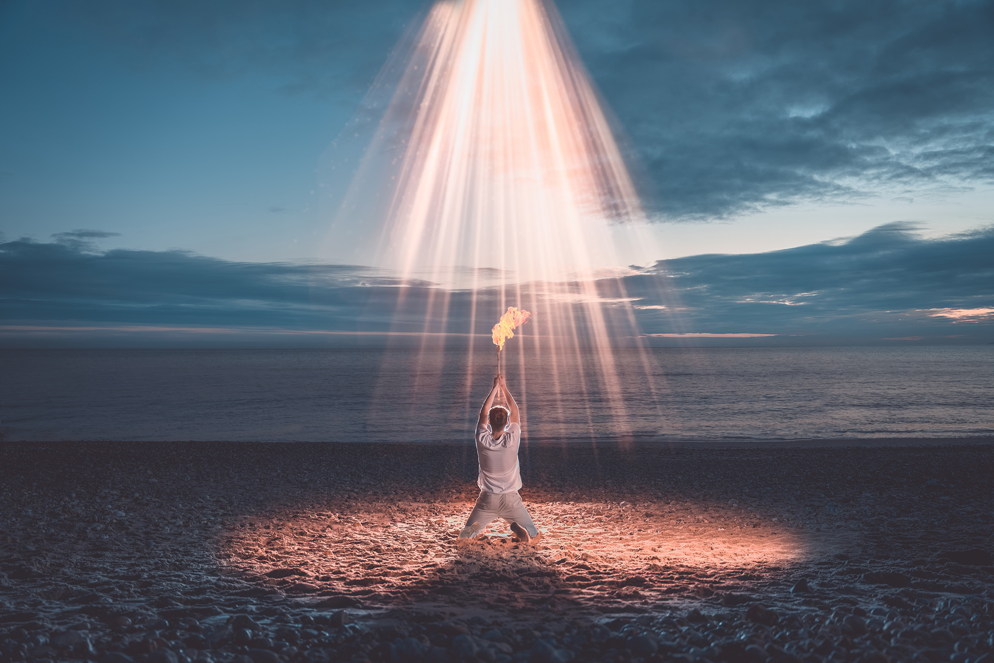 Man kneeling on beach at sunset, holding a flaming torch above his head as a light shines down on him from the sky.