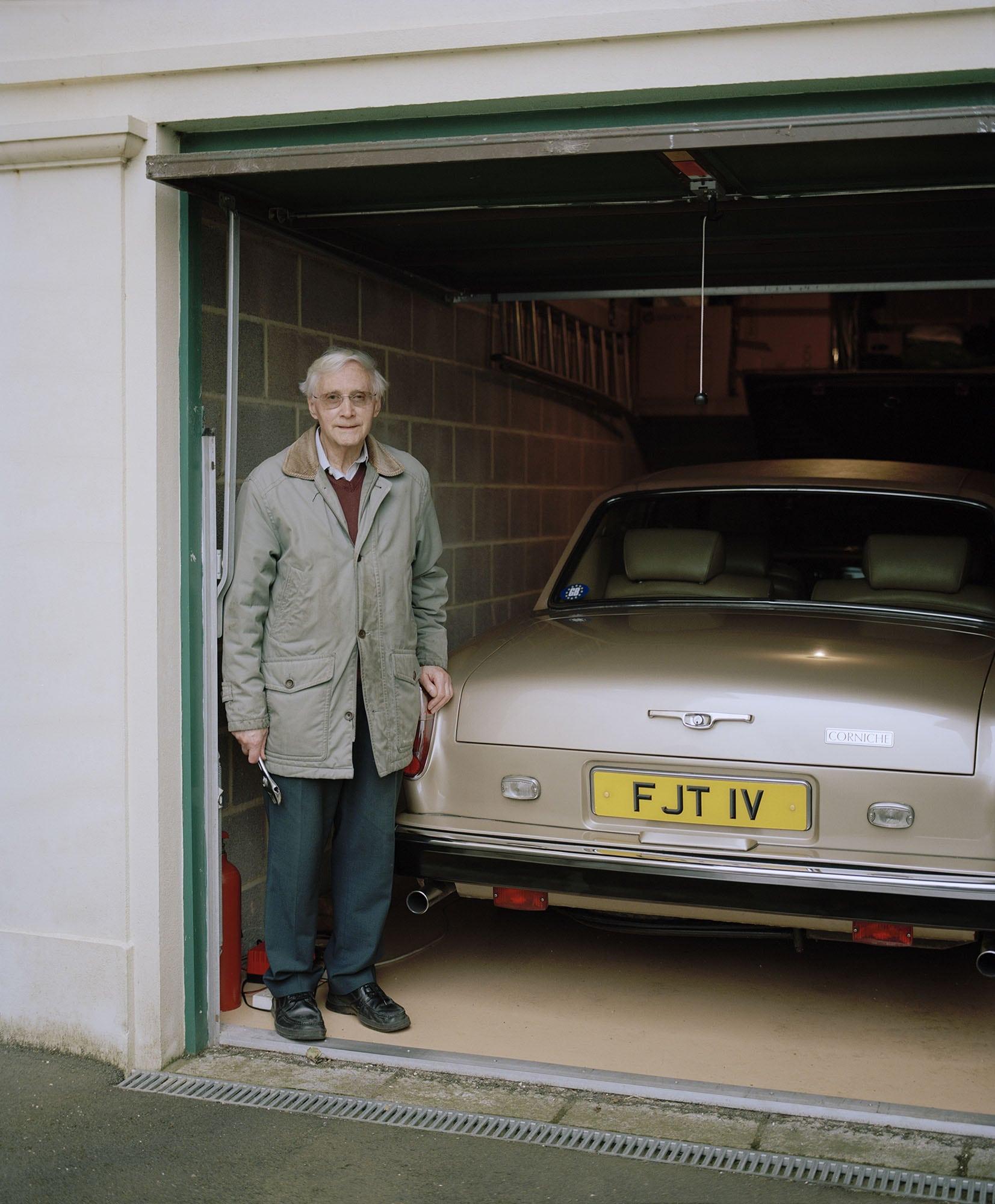 A man stands in a garage doorway next to vintage car with a spanner in his hand.