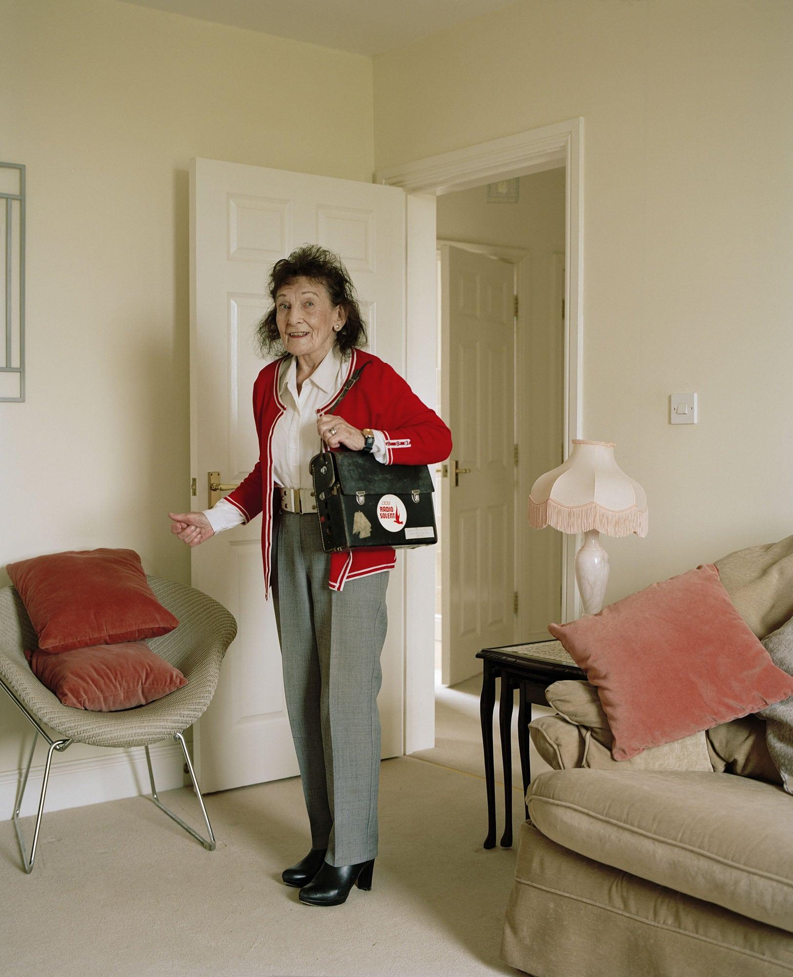 An older lady stands in a living room with a handbag over her shoulder. There is a chair with two cushions to her right and a table, a lamp and a sofa to her left.
