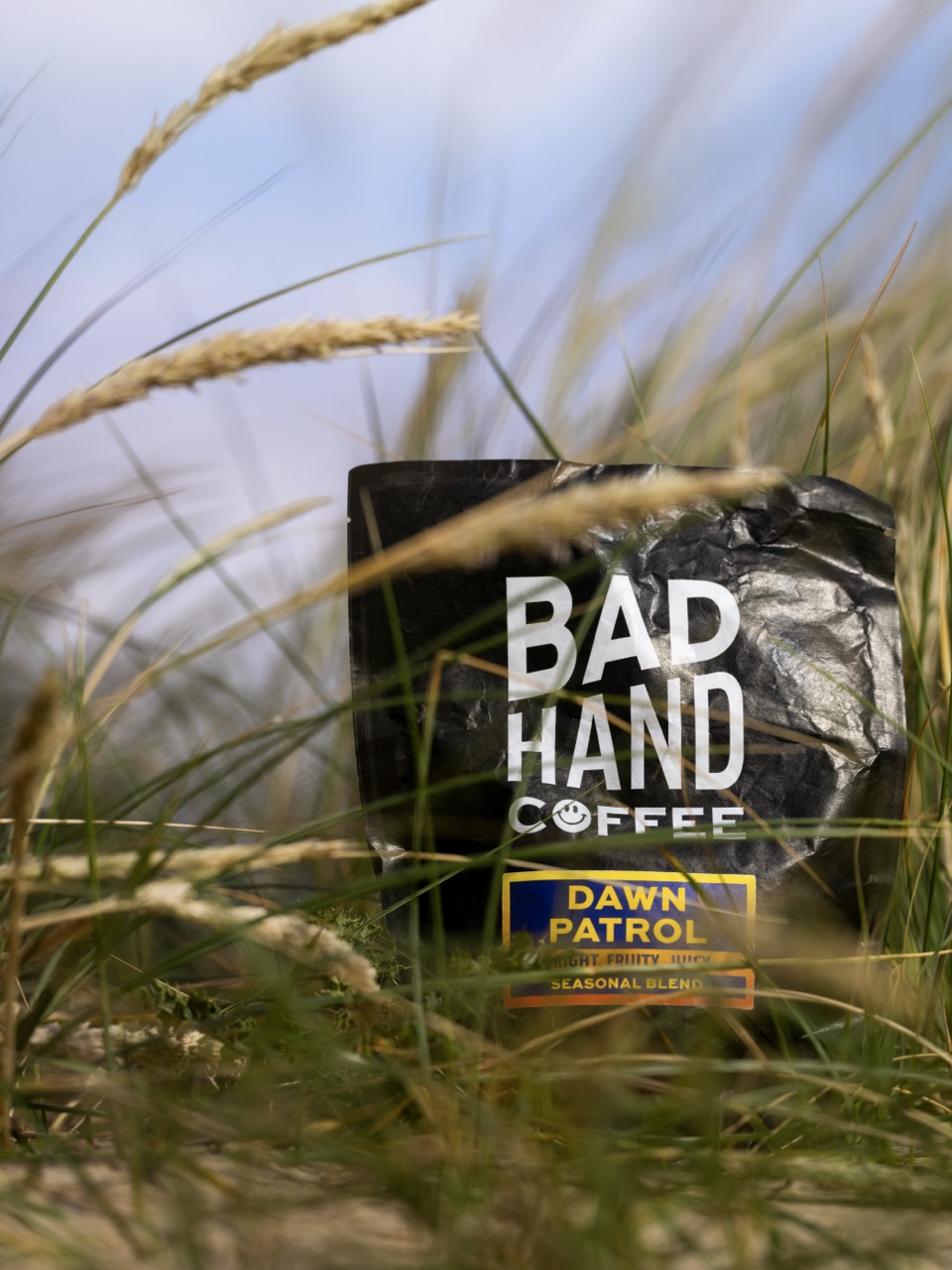 A black coffee bag labelled 'BAD HAND COFFEE' with the subtitle 'DAWN PATROL' sits among tall grasses under a blue sky. The bag features a simple design with bold white text.