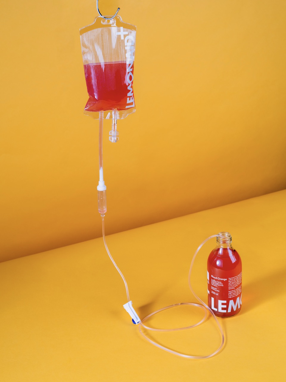 An intravenous drip bag filled with a red liquid hangs from a metal stand, connected by a clear tube to a small red bottle labelled "LEMON". The background is a solid yellow.