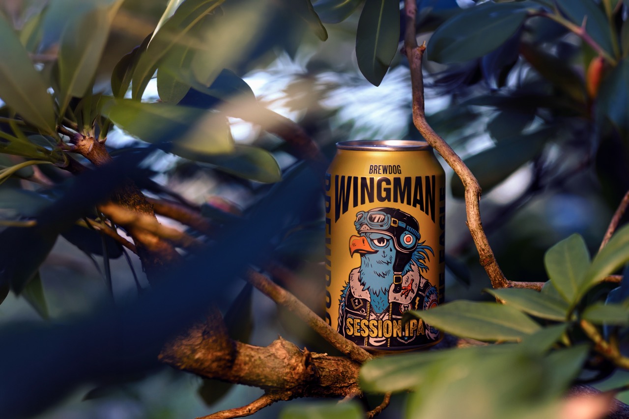 A can of BrewDog Wingman Session IPA is positioned on a tree branch, surrounded by green leaves. The can features a colourful design with a bird wearing sunglasses and a headset.