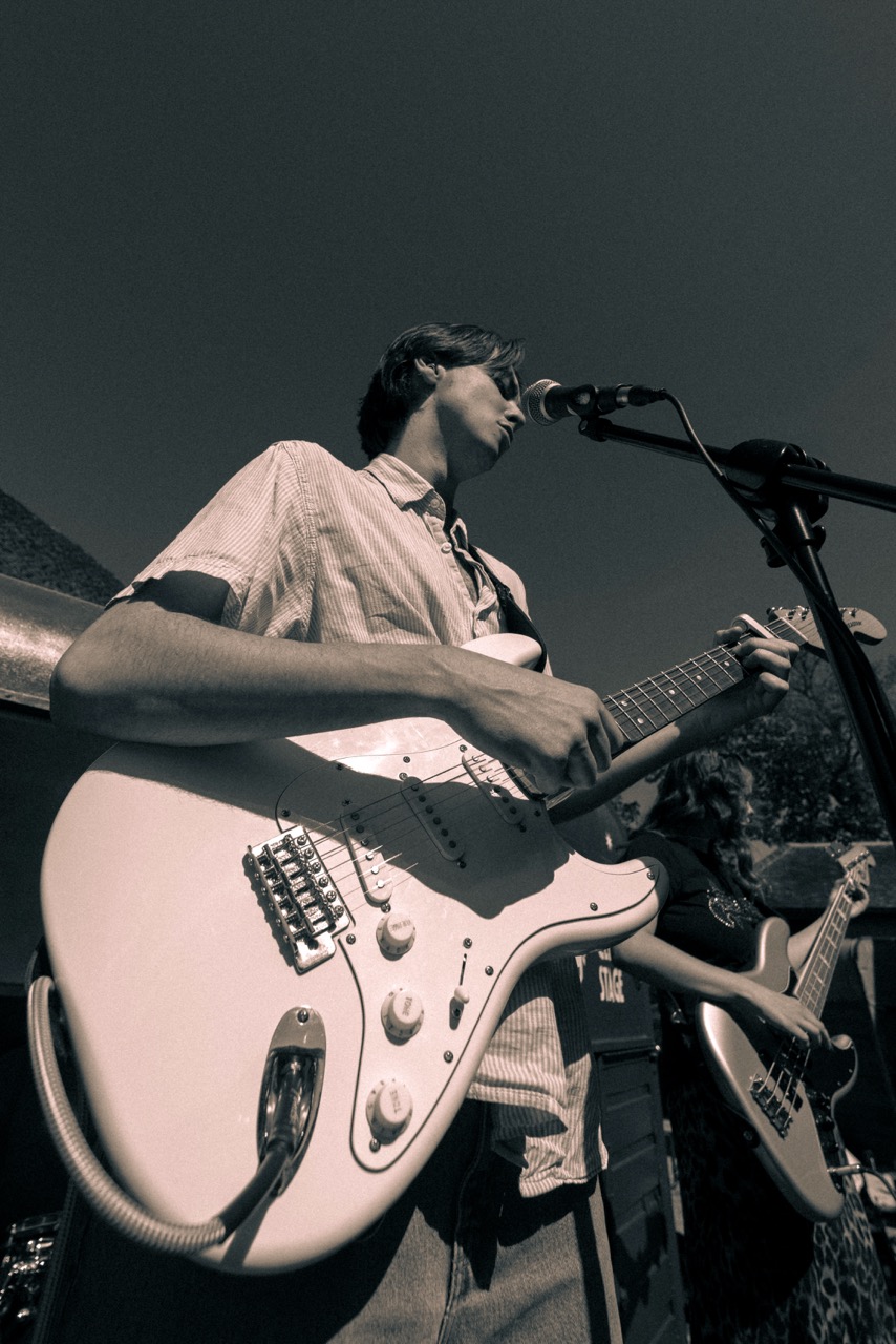 A young man playing an electric guitar on stage, wearing a light-coloured shirt. He is positioned in profile, singing into a microphone, with another musician visible in the background playing a bass guitar. The photo has a black-and-white filter.
