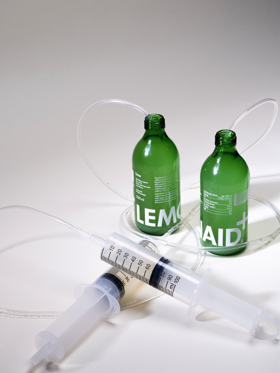 Two green glass bottles labelled "LEMON" and "AID+" are positioned upright, connected by a clear plastic tube. In the foreground, there is a white syringe with visible measurement markings, placed on a light surface.