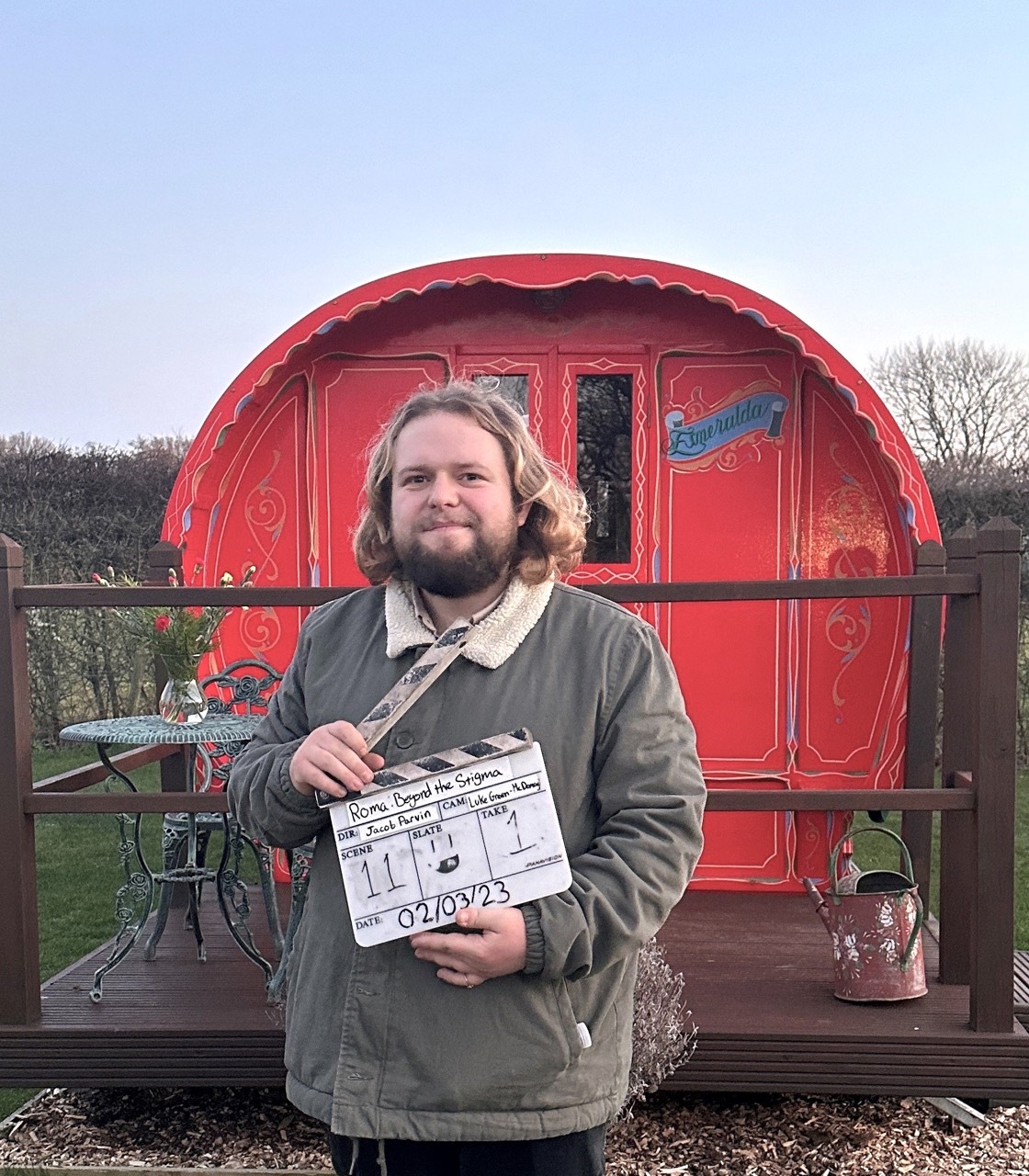Jacob Parvin stood by a traditional Romany home in red, holding a clapperboard.