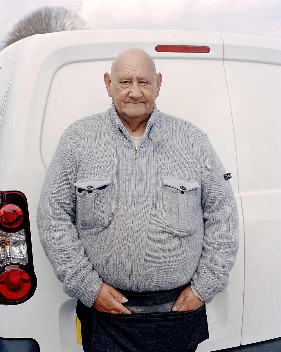 An older person standing with his hands in his apron pockets, the back door of a white van in the background.