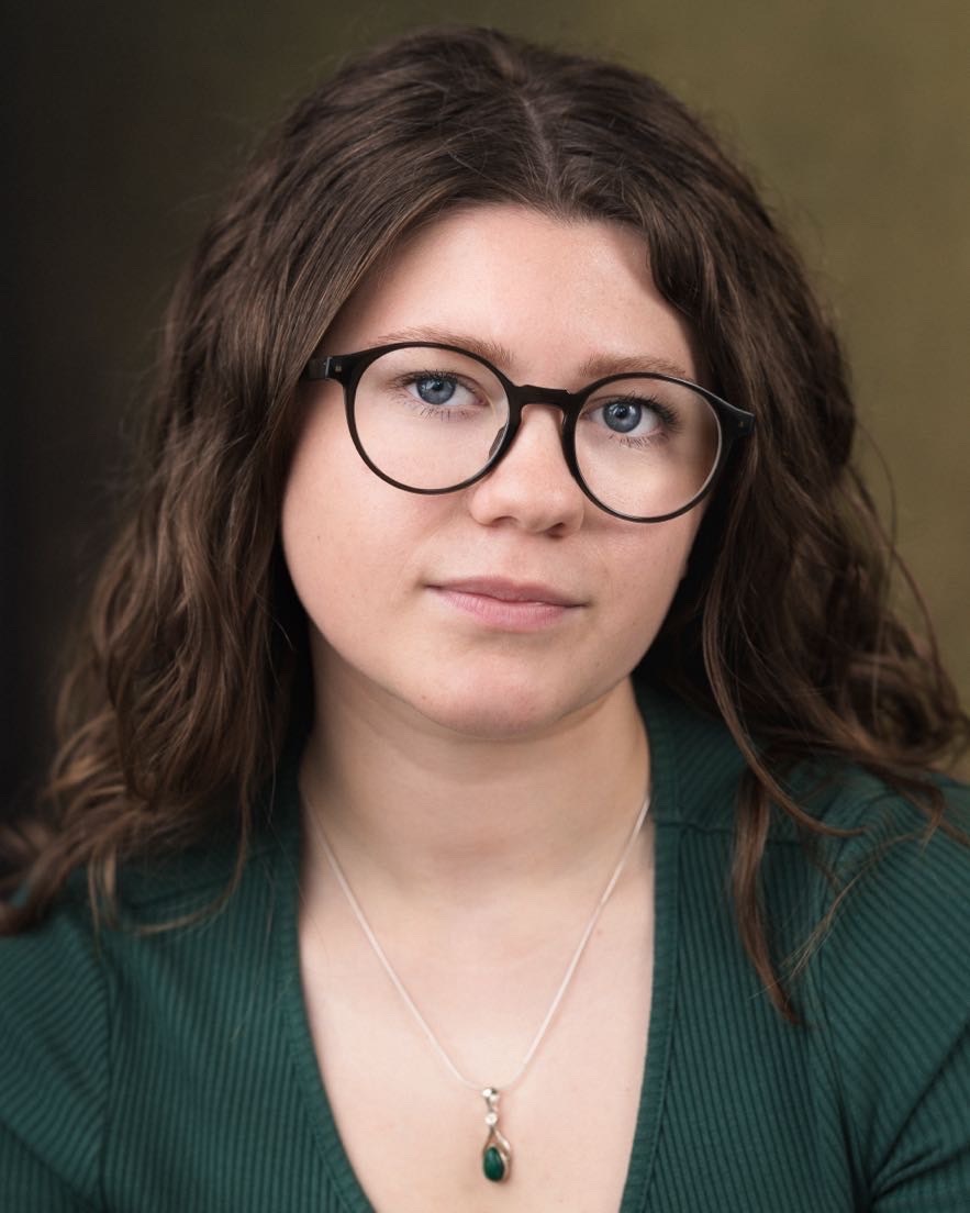 Woman with brown wavy hair wearing black-rimmed glasses and green cardigan with pendant necklace against blurred brown background.