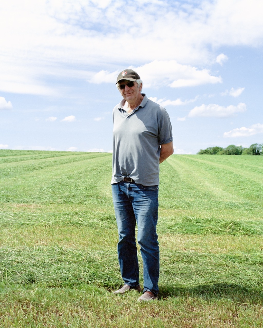 Man in grey polo shirt, cap and jeans standing in green field under cloudy blue sky.