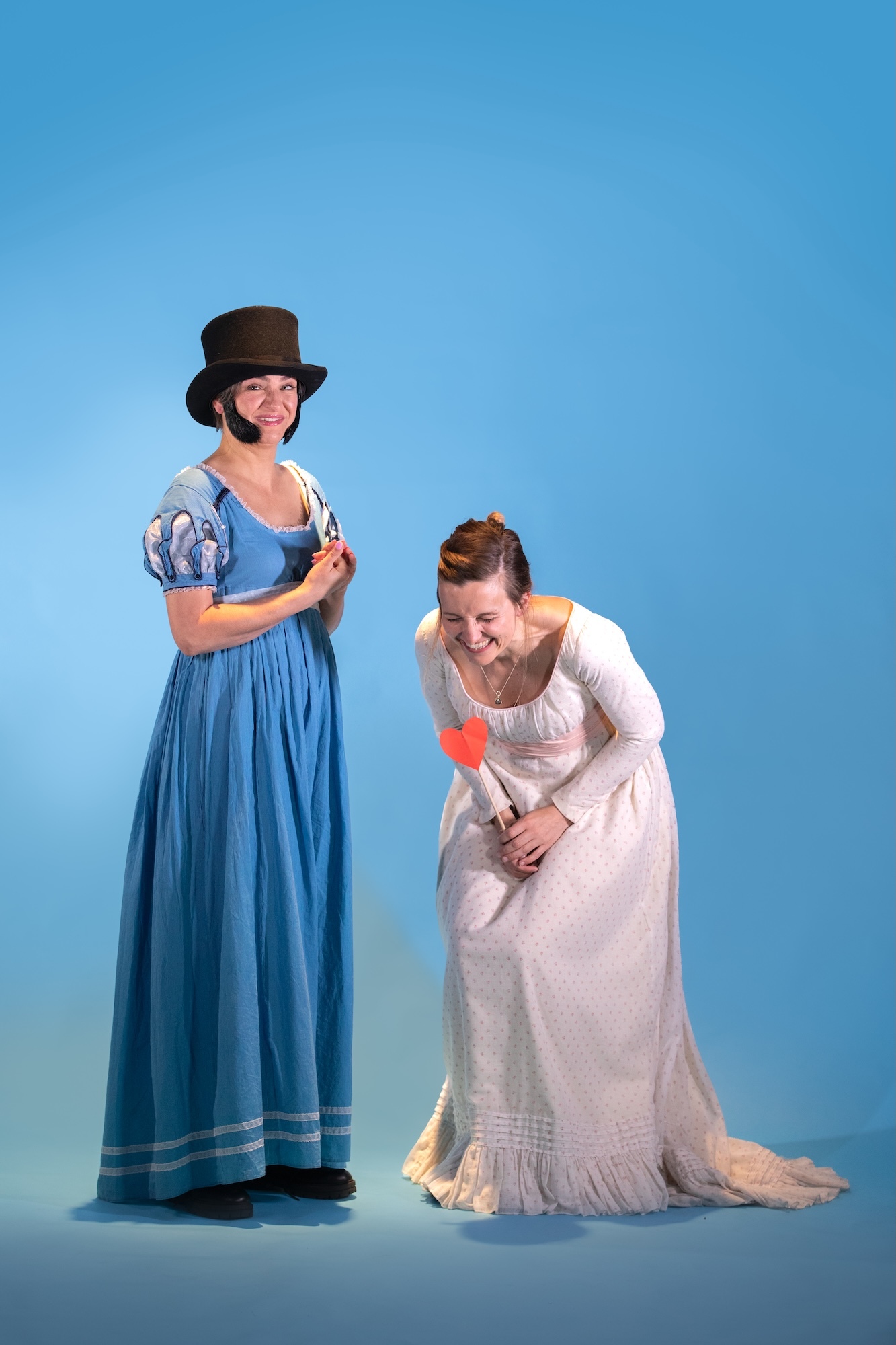 Two people in period costume against blue backdrop. Left figure wears blue dress with white sleeves and dark top hat. Right figure kneels in white dress holding red object.
