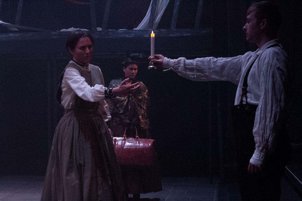 Three actors in period costume on a stage. One is handing a candle to another.
