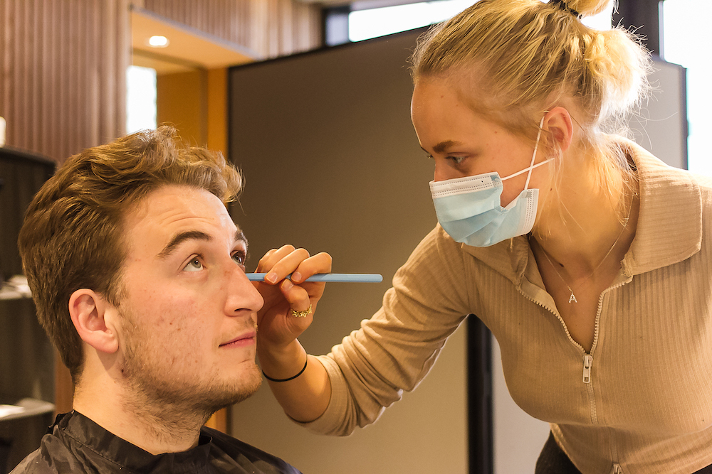 A makeup artist wearing a face mask applies makeup to an actor's face.