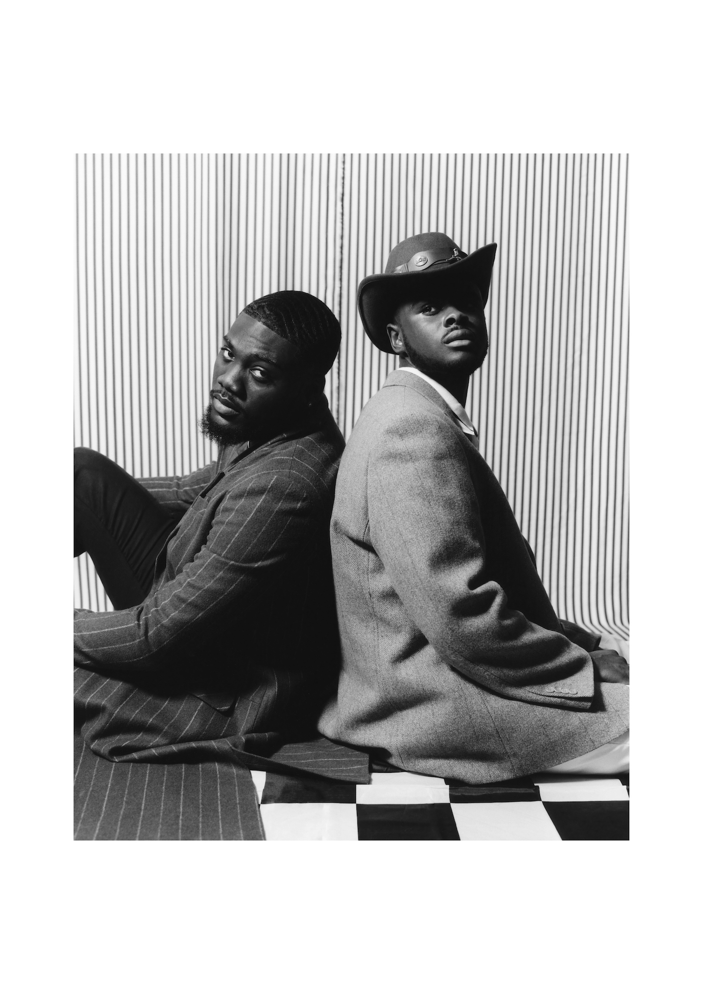 Black and white photo of two models sitting back-to-back on a check-tiled floor, both looking towards the camera.