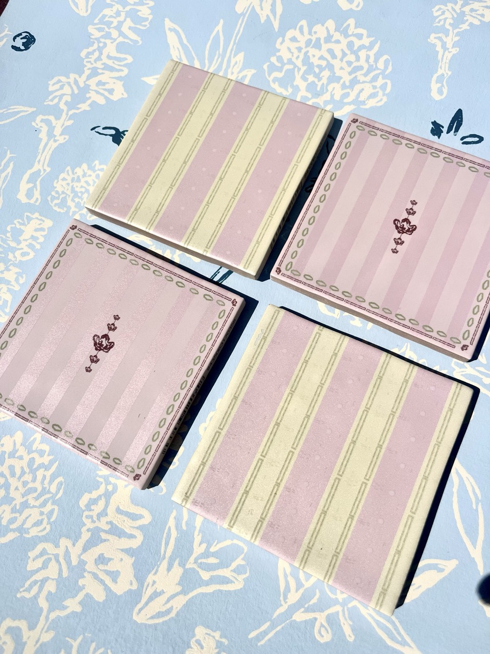 Four square decorative coasters are arranged on a patterned blue surface. Two coasters feature pink stripes and two more have a pink background with floral designs. Each coaster has subtle embellishments along the edges.