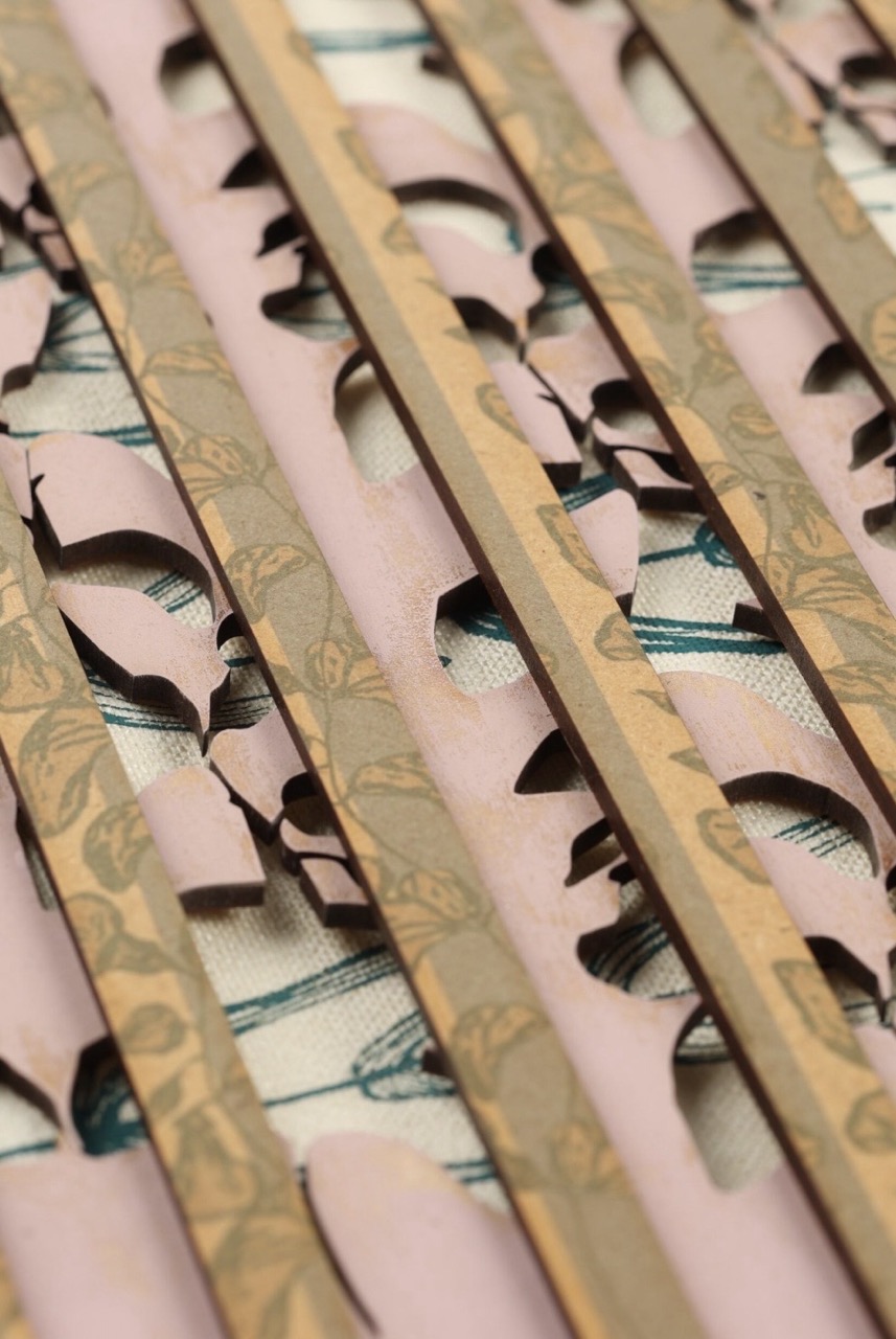 A close-up view of layered wooden strips with intricate cut-out floral patterns, over a soft pink base. The background features a cream fabric with printed designs, partially visible through the cut-outs of the wooden strips.