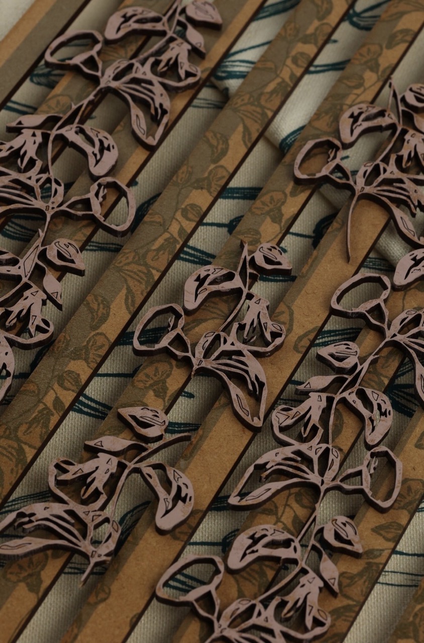 Close-up of patterned brown and cream strips, featuring intricate cut-out floral designs. The background reveals a subtle leaf print in soft, muted tones, enhancing the overall texture.