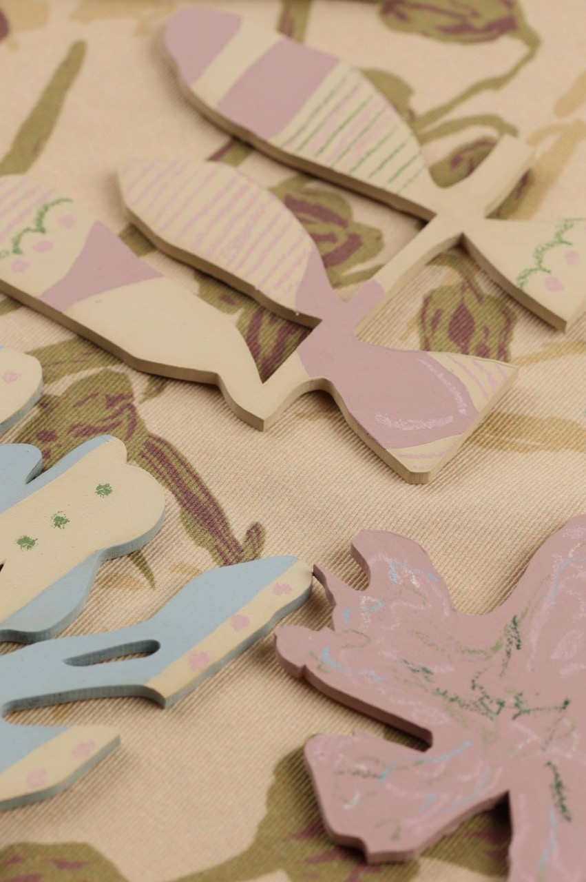 Decorative wooden shapes in pastel colours, including fish and leaf designs, are scattered on a floral fabric background. The shapes feature various patterns like stripes and dots.