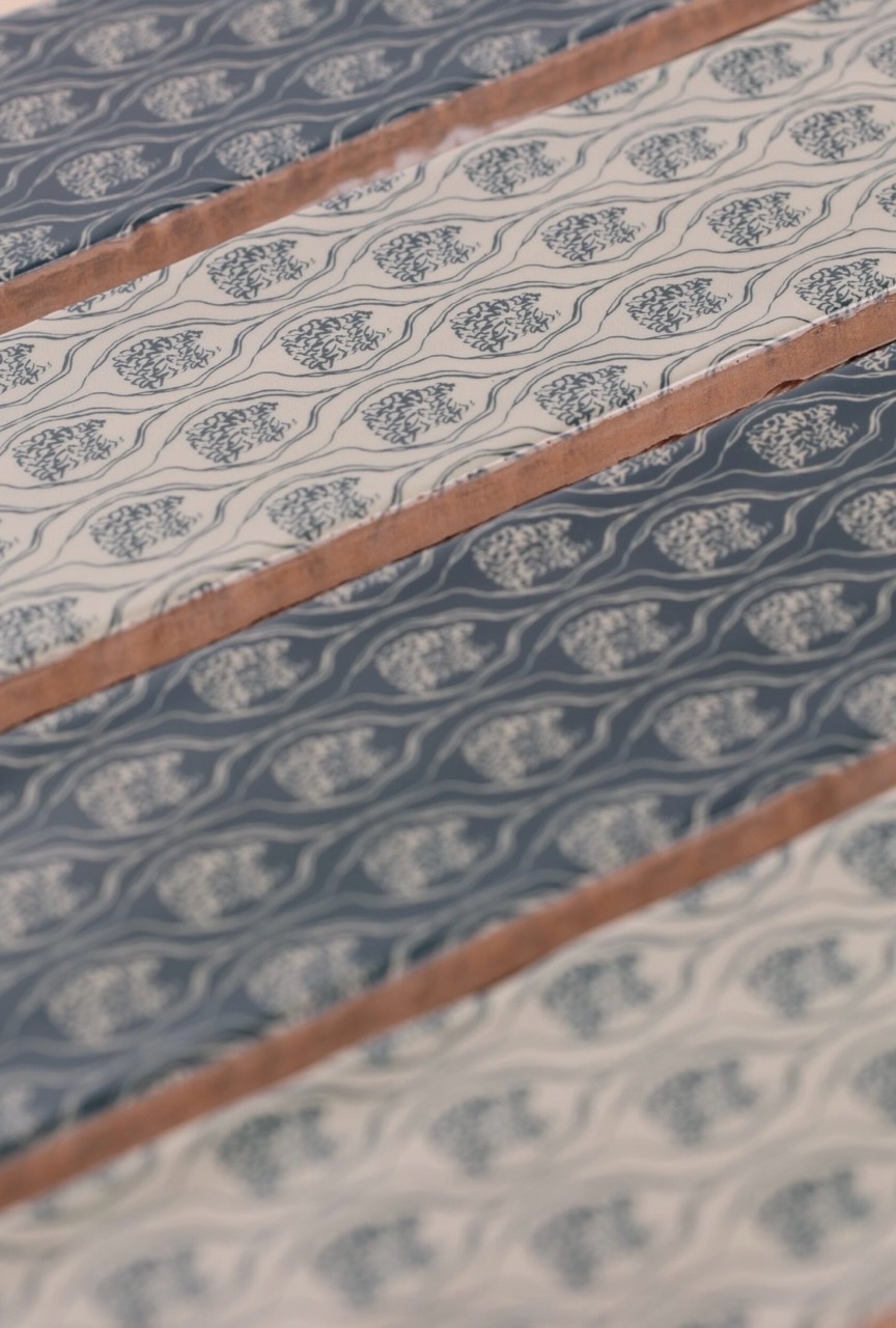 A close-up view of fabric featuring a repeating floral and leaf pattern in shades of blue and cream, separated by rows of light brown satin ribbons.