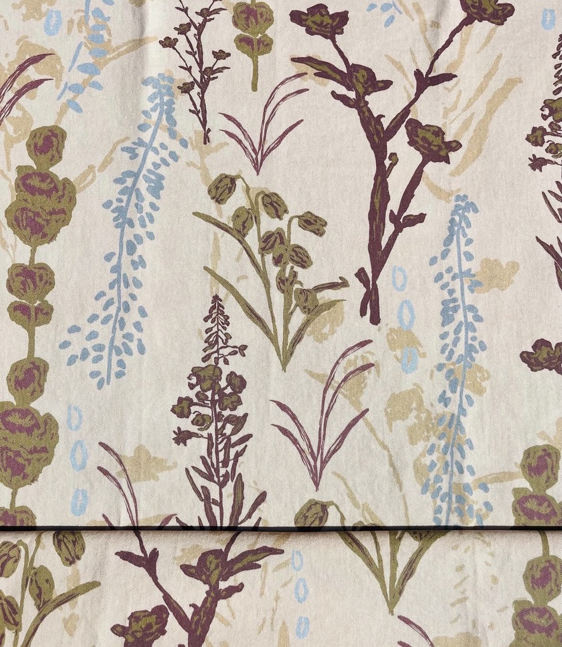 A patterned fabric featuring various floral designs in muted shades of purple, green, and blue against a light beige background. The floral motifs are artistically arranged, creating a natural, organic look.
