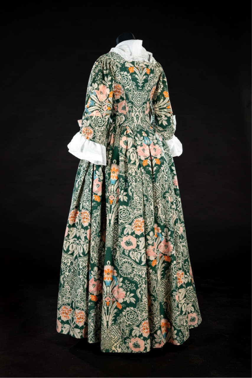 18th century gown with green floral brocade fabric, white lace collar and cuffs, full skirt, displayed against black background.