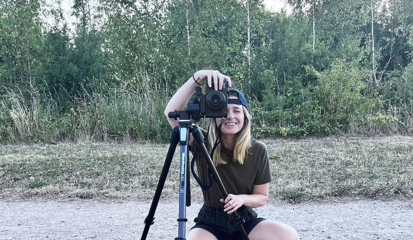 Woman kneeling beside camera on tripod, smiling at viewer with dense green trees and scrubland in background.