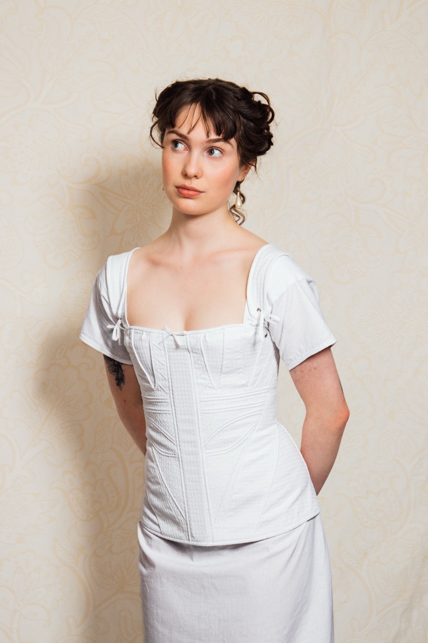 Katie is wearing a regency period corset and chemise.The corset is corded and embroidered, and features a little bow decoration at the neckline.