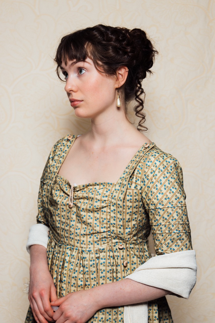 Katie is wearing a regency period empire line dress. The dress has a ‘bib front’ opening, which buttons at the shoulders. The dress is a straw brown colour, and features a blue and brown floral check pattern. She is wearing a cream-coloured shawl around her elbows.