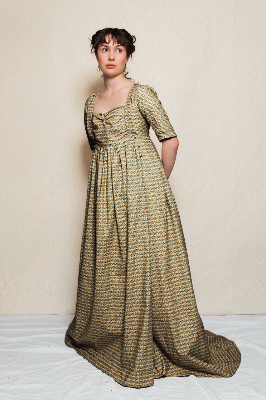 Katie is wearing a regency period empire line dress, with a train. The image shows the full length of the dress. It features a high waist and gathered skirt.
