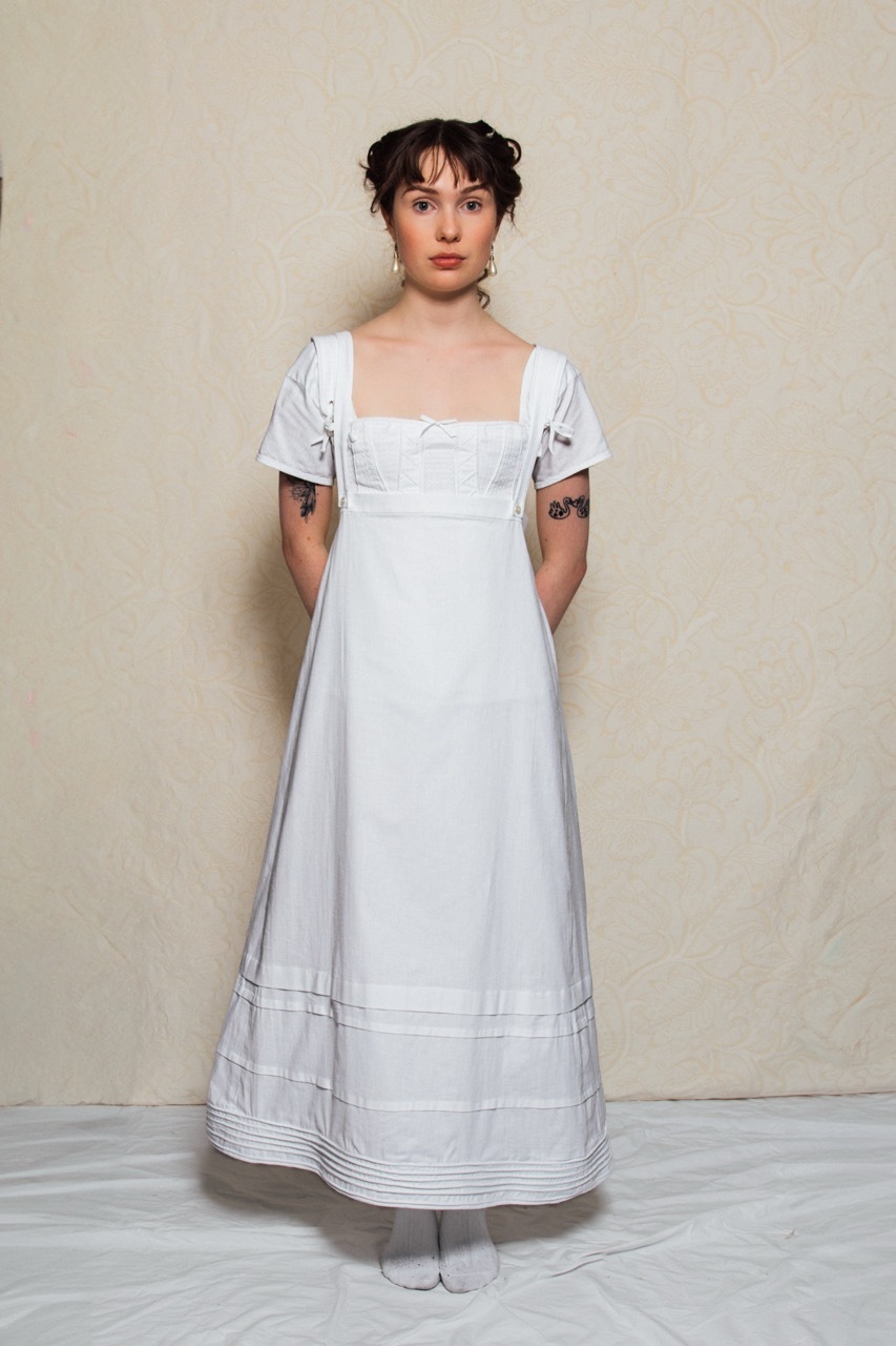 Katie is wearing regency period undergarments. This includes a chemise, corset and petticoat. The petticoat features horizontal pleats and cording at the hem. The petticoat is held up by two straps over the shoulders.