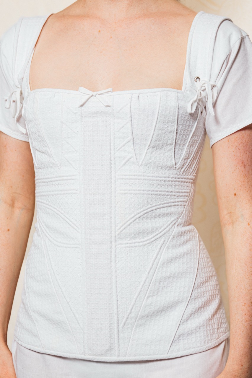 A close-up of the details on the corset. The corset is corded and embroidered, and features an inch wide wooden busk down the centre-front. The corset fits over the hips, and is fastened with lace and eyelets at the back.