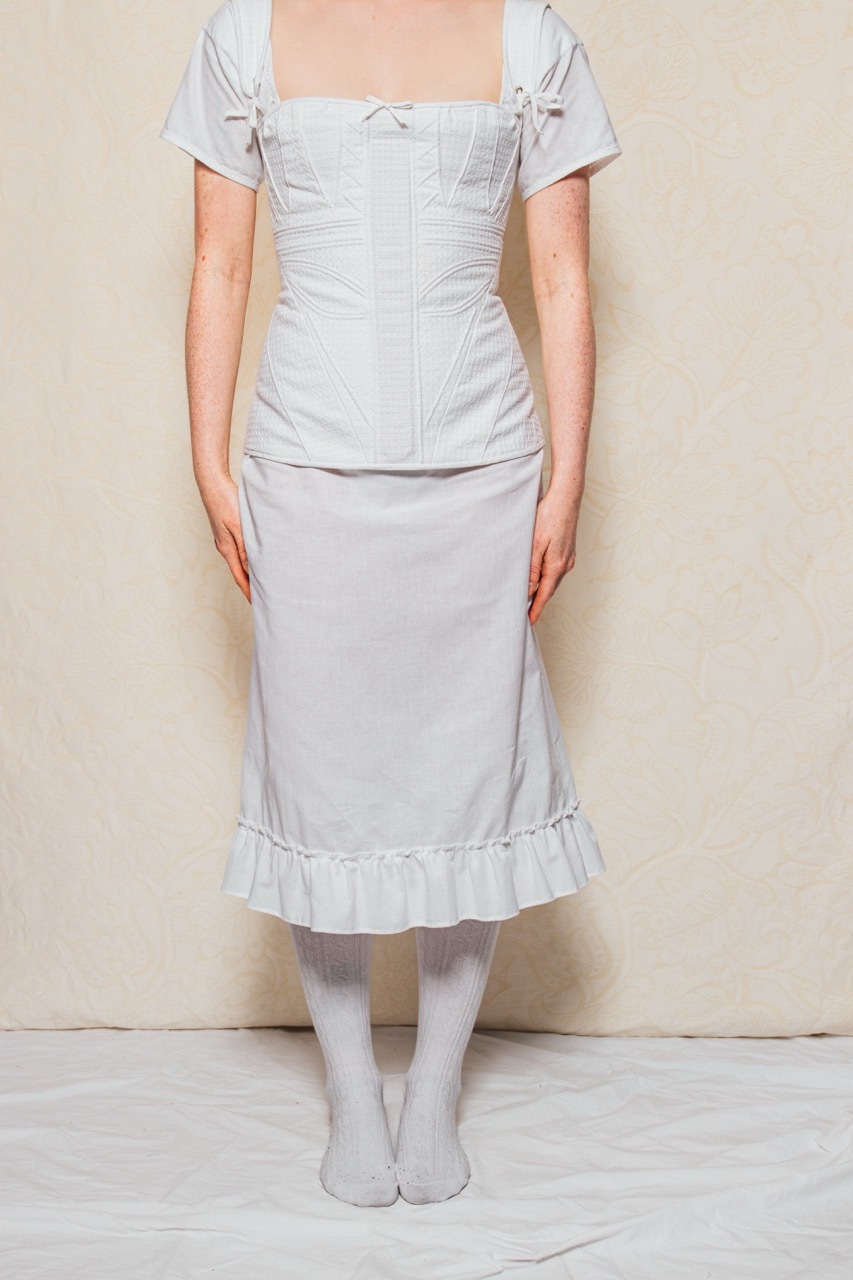 A full view of the corset and chemise. The chemise, worn underneath the corset, features a gathered frill at the hem.
