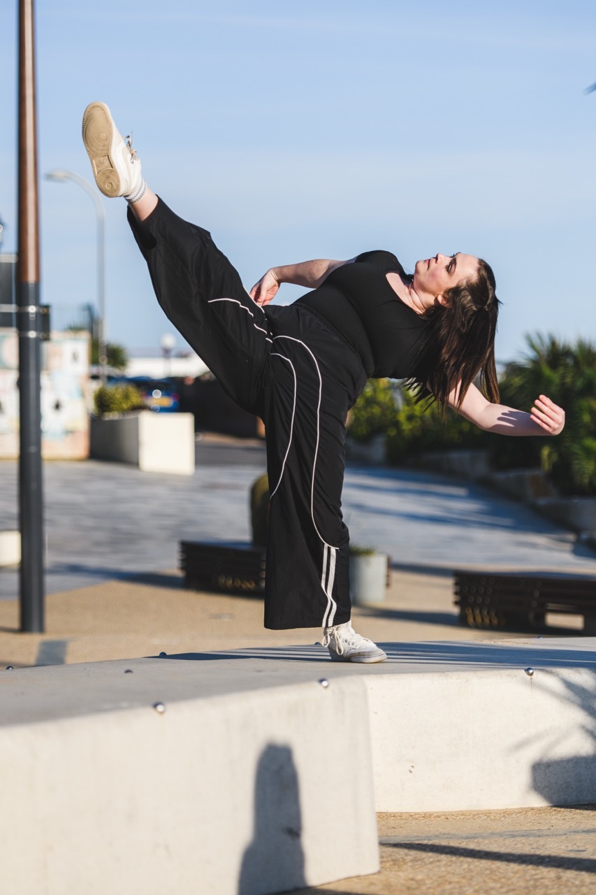 Woman in black tracksuit performing high kick on rooftop terrace with blue sky and urban buildings in background.