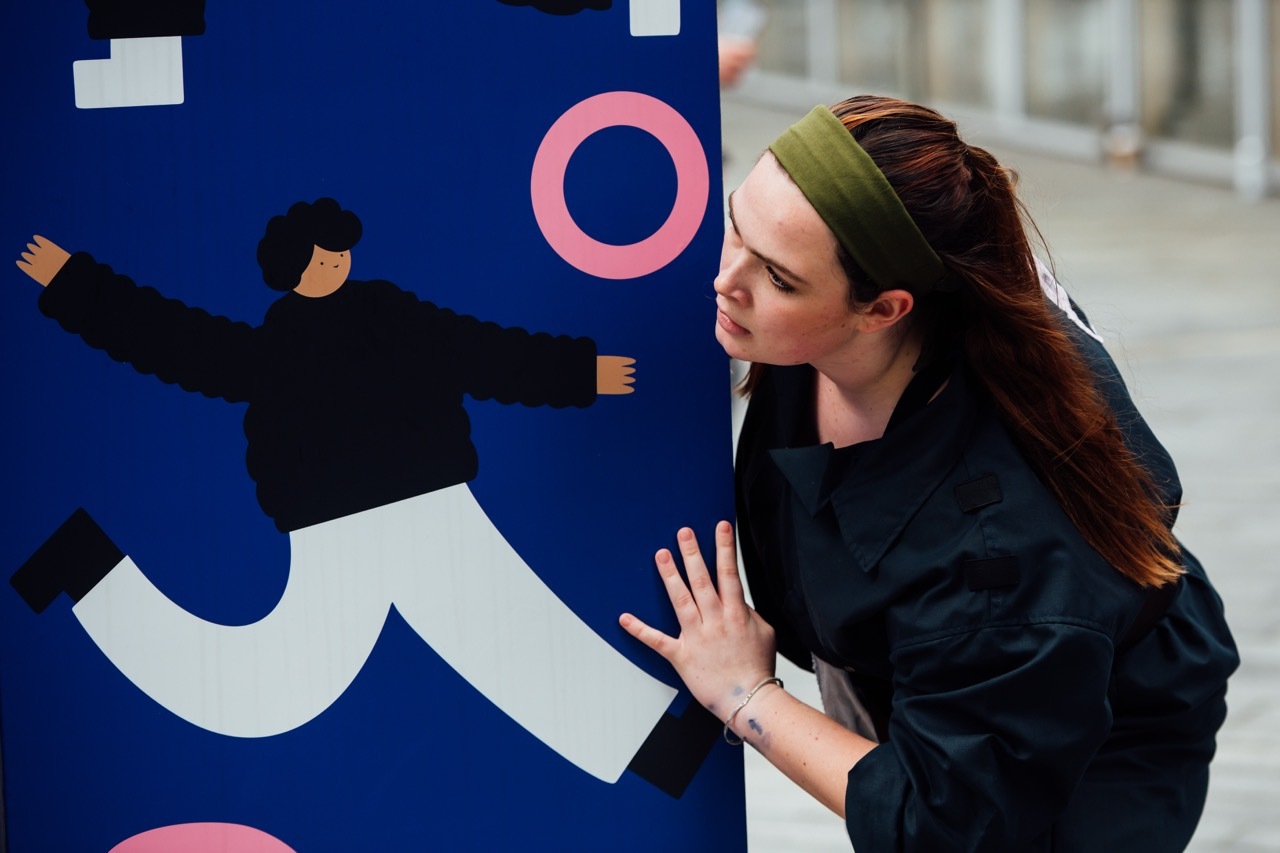 Woman with green headband and dark clothes poses with hand raised against blue wall, featuring illustrated person, white wavy design and pink circles.