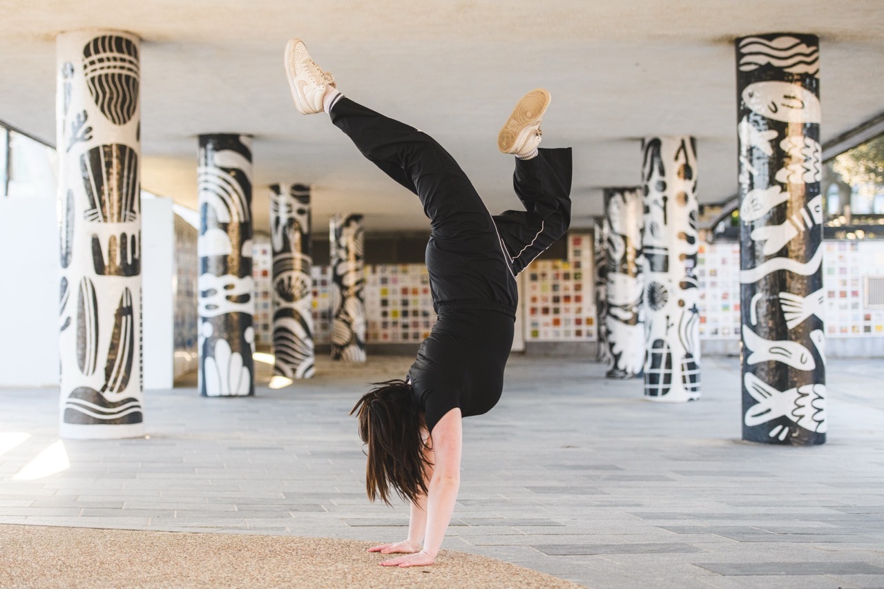 Woman performing handstand in black workout clothes inside studio with decorative patterned columns.
