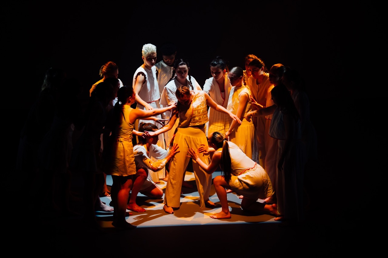 Theatre performers in white and orange costumes clustered together under warm stage lighting against black background.