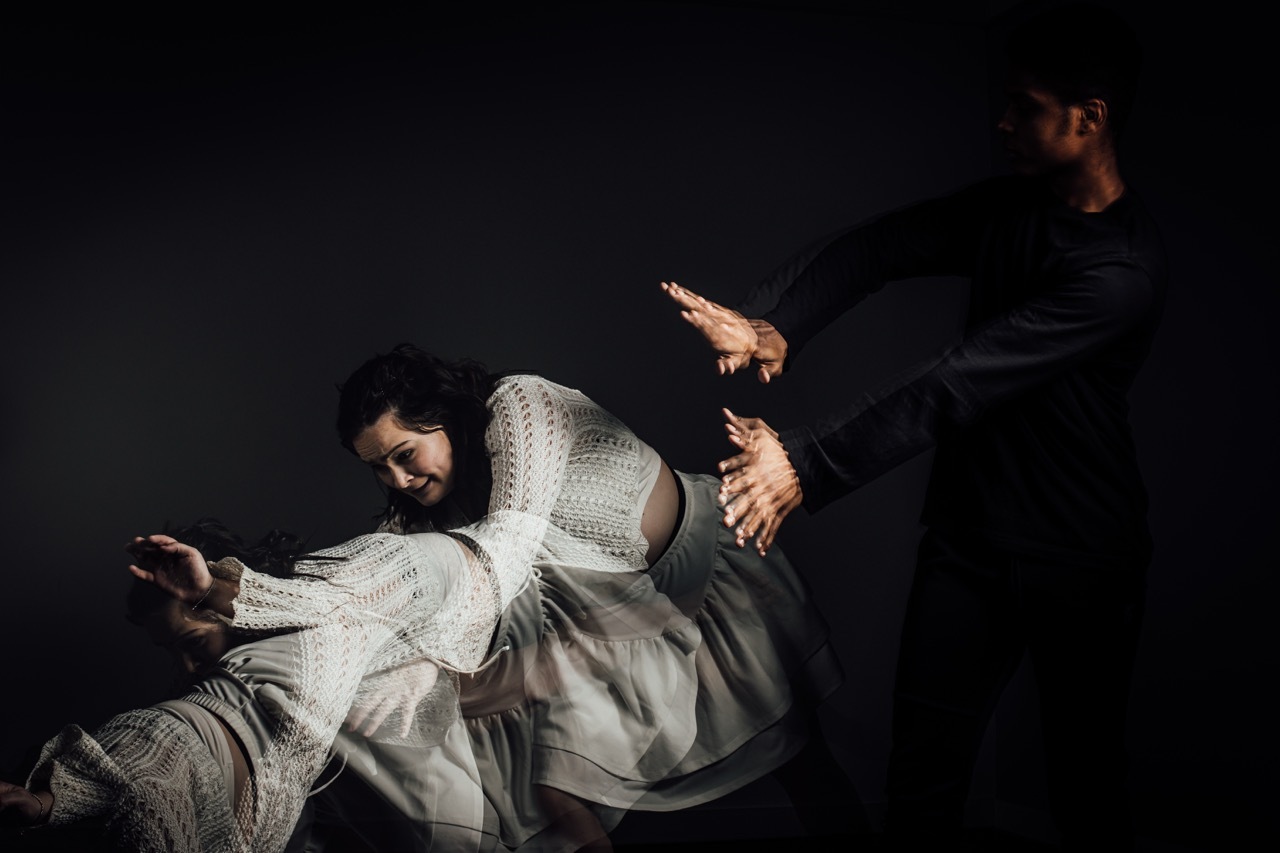 Two figures against dark background: person in white dress seated on left, figure in dark clothing standing on right with outstretched arms.