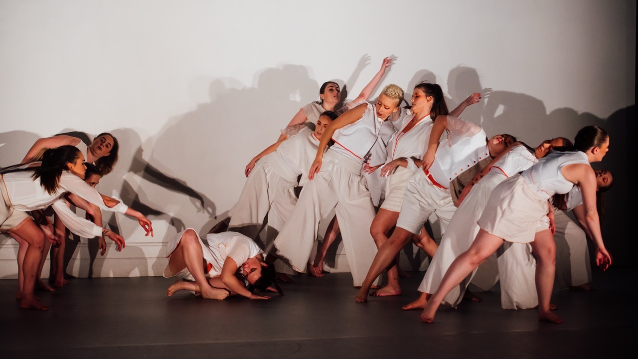 Group of dancers in white costumes with red accents performing on dark stage, bodies angled and arms extended in dynamic poses.