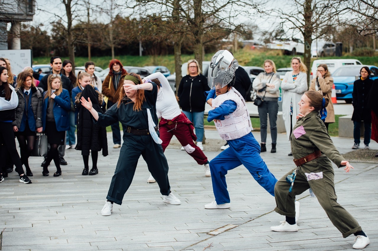 Group of people dancing on paved surface, one in blue with a large metal mask, spectators watching in background, bare trees and buildings visible.