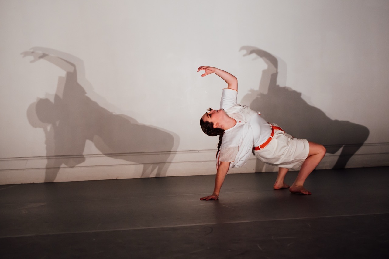 Person in white clothing performing synchronised horizontal movements against a white wall, casting dramatic grey shadows.