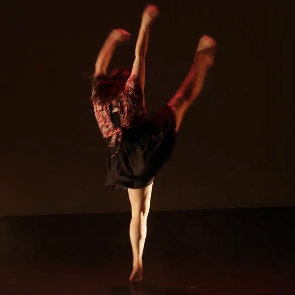 Dancer in red top and dark skirt performing with arms raised and one leg extended, against warm orange-brown lighting.