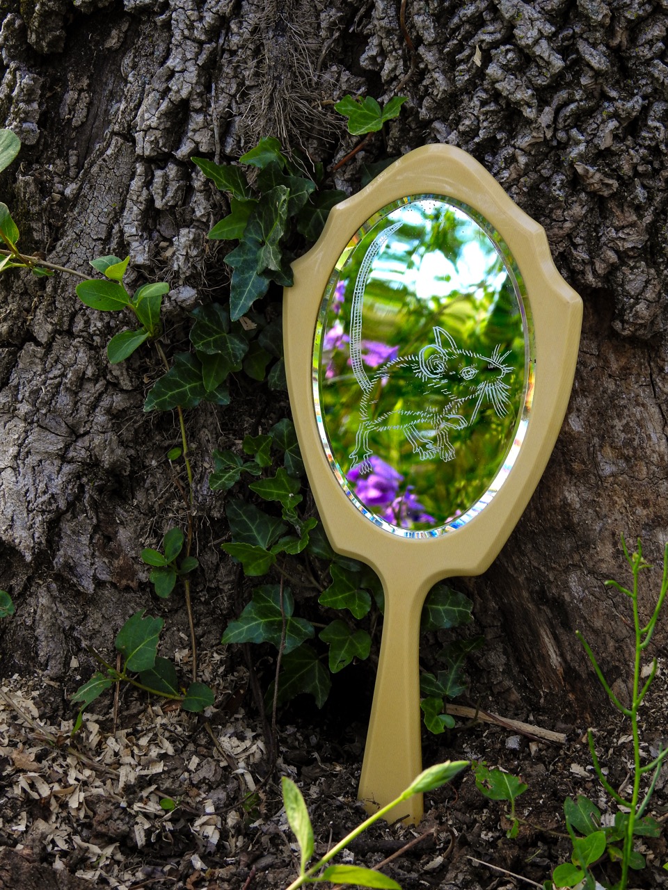 A cream-handled oval garden mirror leans against a tree trunk, reflecting green foliage and purple flowers amid ivy and bark.