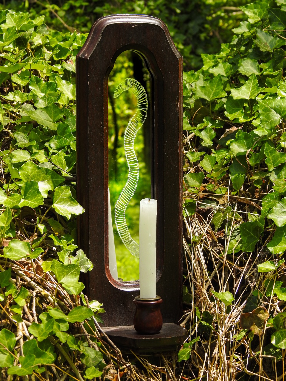 A white candle in a dark wooden memorial frame stands among dense ivy, with a green spiral glass ornament reflected in the narrow mirror panel.