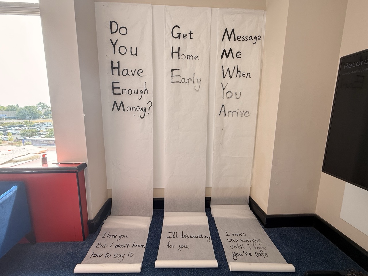Three long paper banners in a room by a window ask “Do you have enough money?”, “Get home early” and “Message me when you arrive”, with smaller handwritten notes at the bottom.