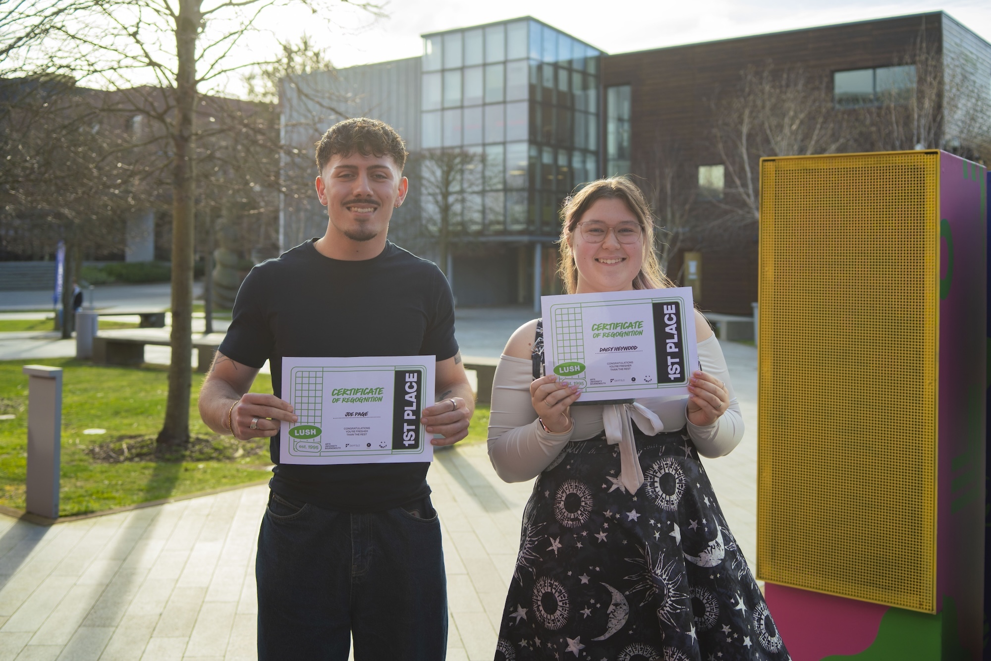 J Page and D Heywood holding certificates stand outdoors beside yellow structure, modern glass building and trees in background.